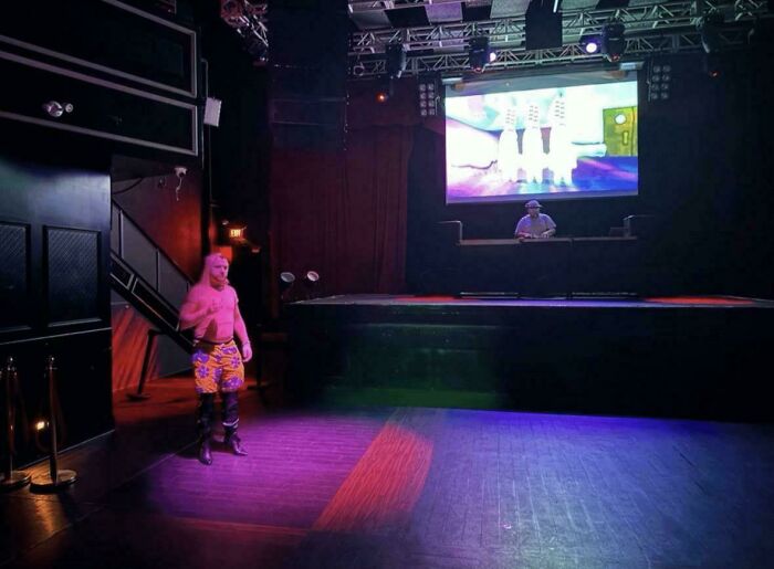 Hombre en un escenario oscuro, vestido con pantalones cortos coloridos, provocando vergüenza ajena.