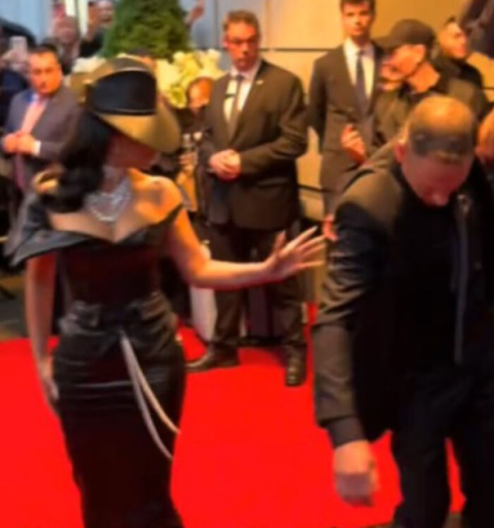 Persona con vestido negro y sombrero en alfombra roja interactuando con hombre en traje, momentos incómodos Gala Met 2025.