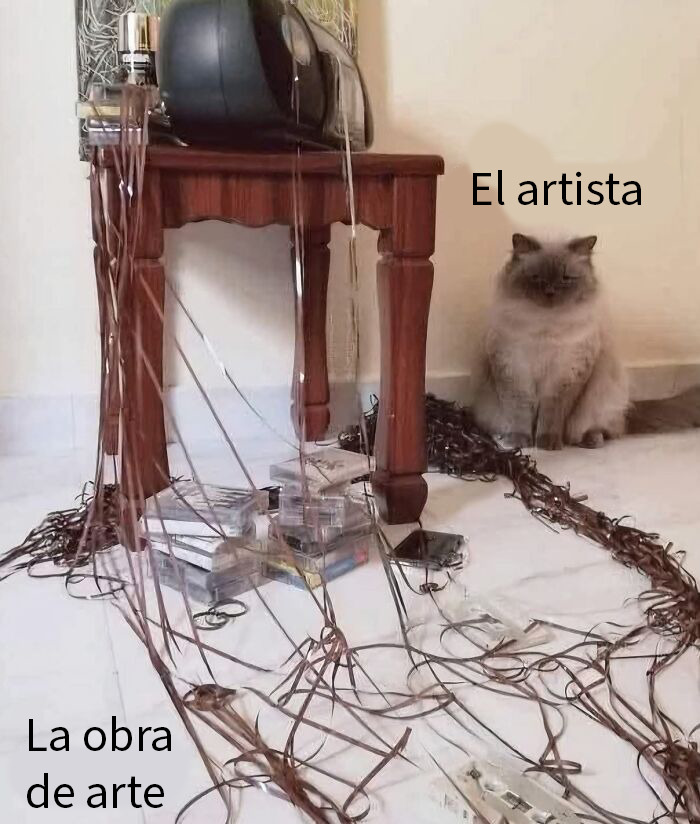 Gato sentado junto a una mesa con cintas de casete desenrolladas, mostrando la mala leche felina en el desastre.
