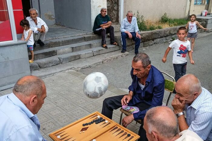 Niños y hombres mayores capturan el momento antes de un desastre con balón de fútbol interrumpiendo juego de mesa.