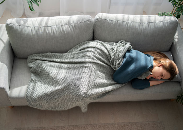 Mujer acostada en sofá cubierta con manta gris, mostrando el cansancio tras historias de drama familiar caótico y divertido.