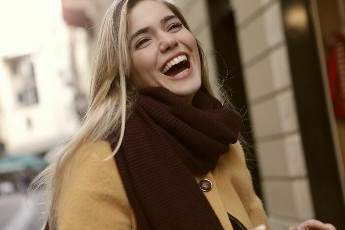 Una mujer rubia sonriendo felizmente en la calle vistiendo un abrigo amarillo y bufanda