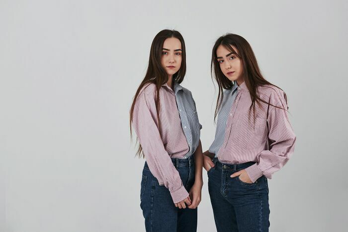 Dos jóvenes gemelas idénticas posando juntas con camisas similares sobre fondo gris.
