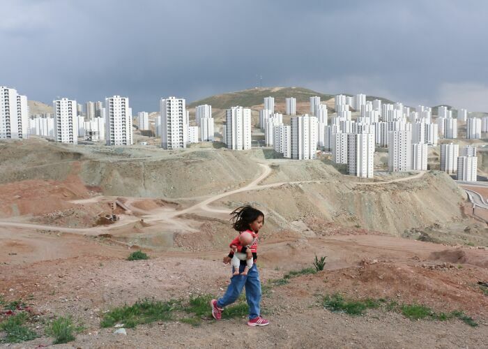 Niña con muñeca camina en terreno árido frente a edificios en los Premios internacionales de fotografía Siena