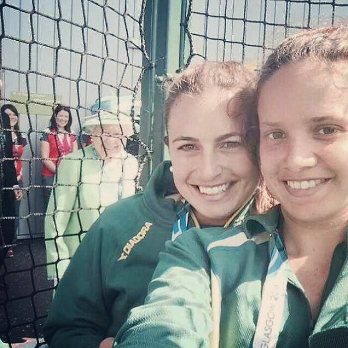 Divertidas fotos con la Reina Isabel sonriendo en el fondo detrás de dos jóvenes en primer plano.