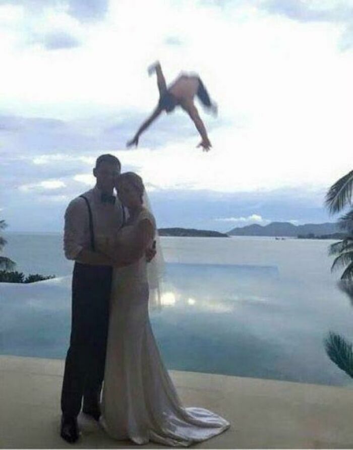 Novios posando junto al mar mientras alguien salta de cabeza al fondo de divertidas fotos de boda.