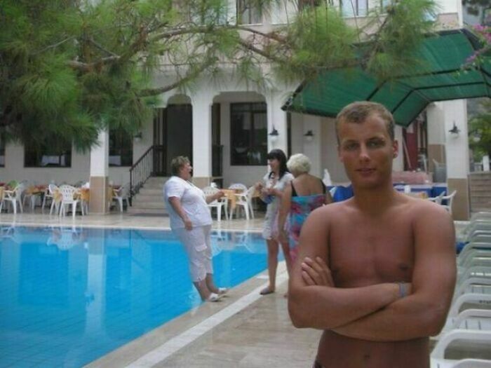 Hombre posando en la piscina mientras una mujer cae al agua de fondo, creando una de las mas divertidas fotos.