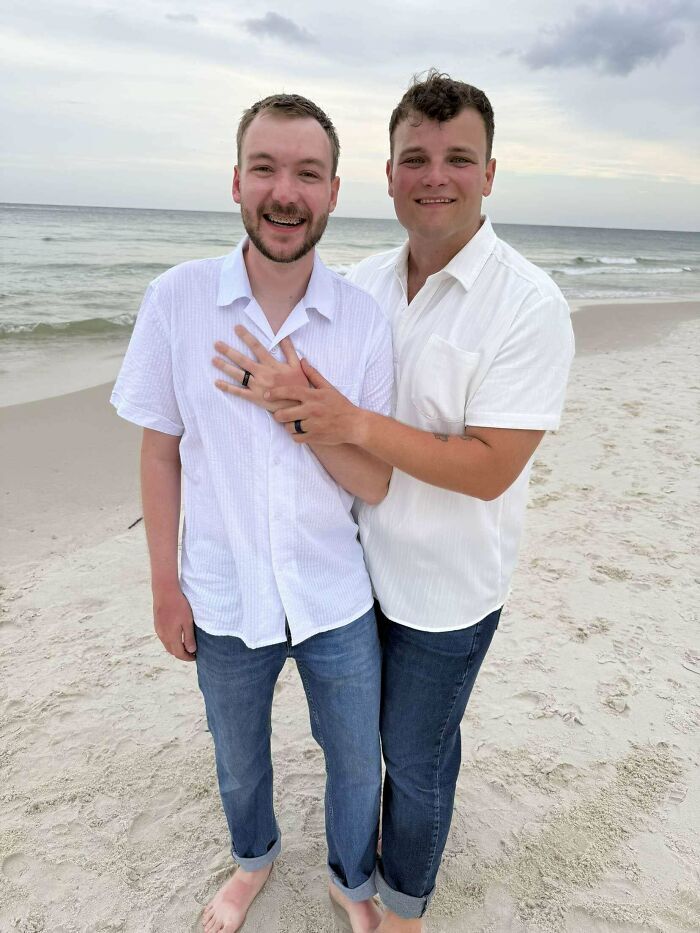 Pareja sonriente abrazada en la playa mostrando sus anillos, una de las publicaciones reconfortantes del día.